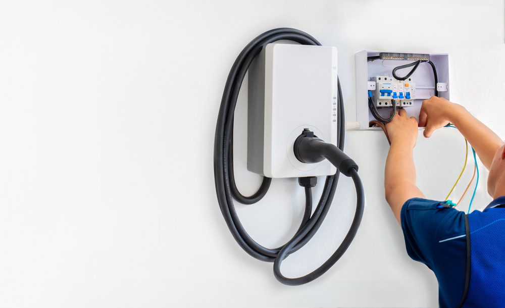 Person installing or wiring an electric vehicle charging station on a white wall, with cables connected to a control box.