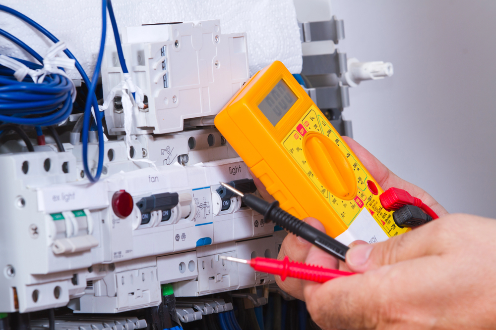 A person uses a digital multimeter to check an electrical circuit inside a control panel with wires, switches, and breakers.