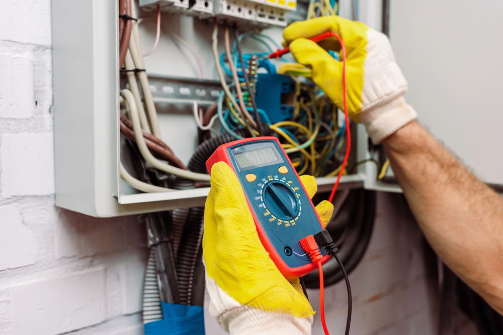Person wearing yellow gloves uses a digital multimeter to test electrical wiring inside an open electrical panel.