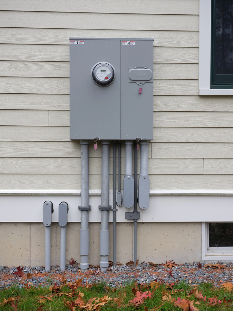 Gray electrical meter and service panel mounted on the exterior wall of a house, with multiple conduit pipes running vertically below.