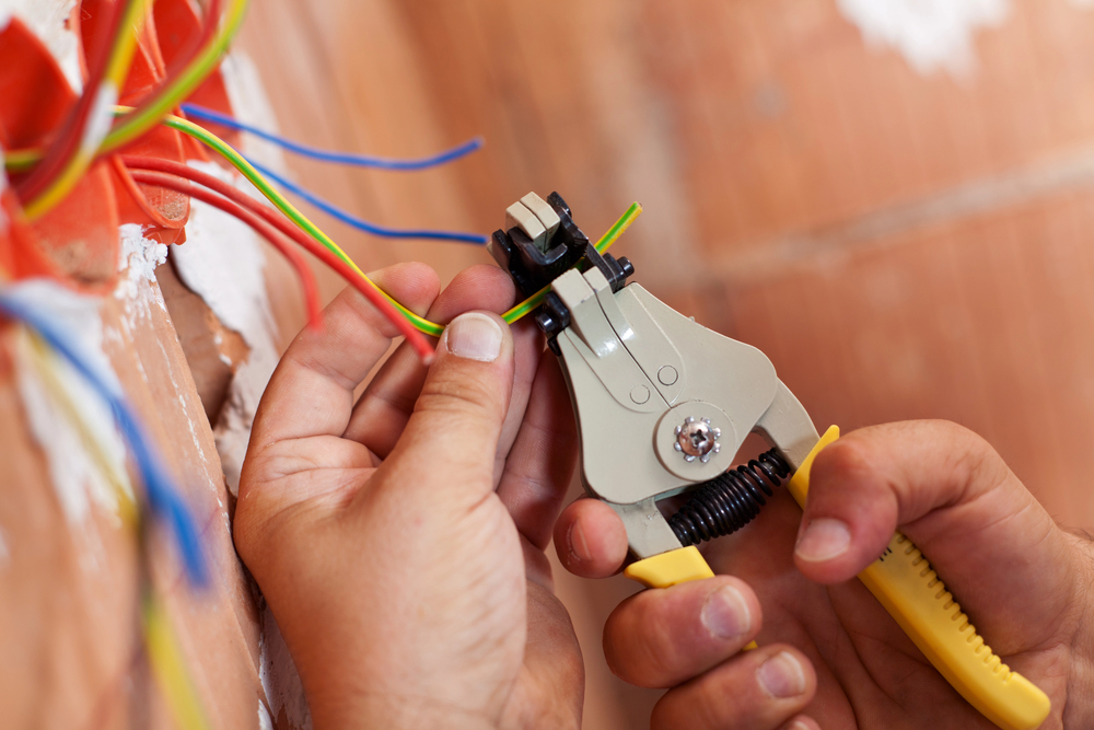 Close-up of hands using a wire stripper to remove insulation from electrical wires against a partially finished wall.