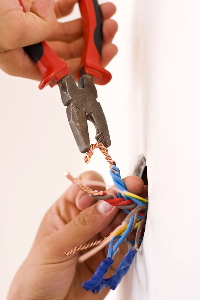 A person uses pliers to cut exposed copper wires extending from a hole in a wall. Several wires, some with blue tape, are visible.