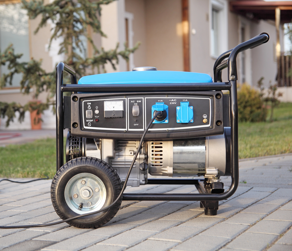 A portable electric generator with wheels and a blue top is placed on a paved area in front of a house.